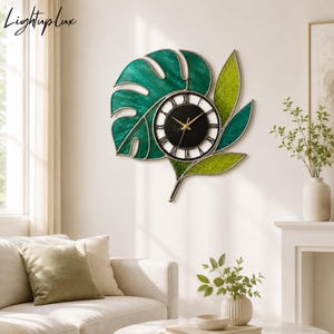 May include: A wall clock featuring a circular black face with white Roman numerals and gold hands, surrounded by a decorative frame shaped like multiple monstera and tropical leaves in varying shades of green, with the text Lightuplux in the corner.