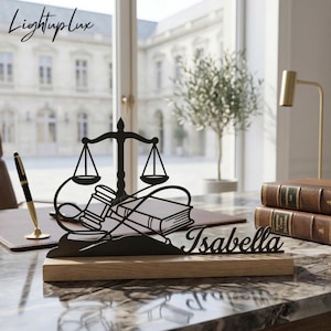 May include: A black metal desk nameplate featuring a scale of justice, gavel, and books design, with the name "Isabella" cut out. The nameplate is mounted on a wooden base. The "LightupLux" logo is in the upper left corner.