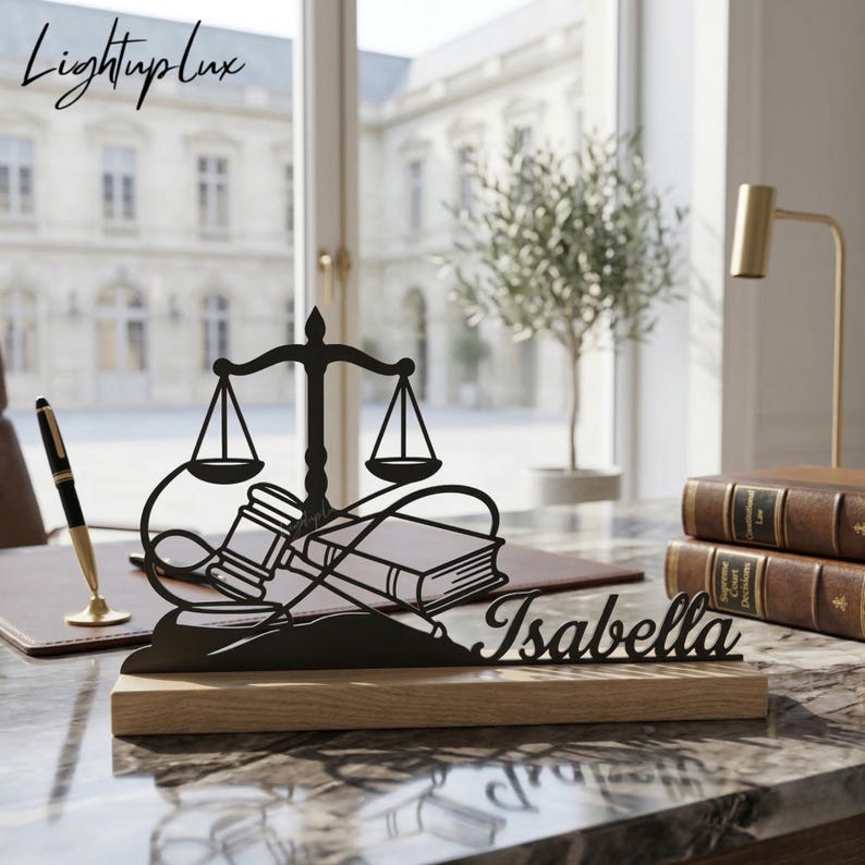 May include: A black metal desk nameplate featuring a scale of justice, gavel, and books design, with the name "Isabella" cut out. The nameplate is mounted on a wooden base. The "LightupLux" logo is in the upper left corner.