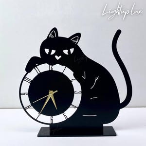 Op de afbeelding: Een zwarte katvormige bureauklok met een ronde wijzerplaat. De kat zit, met zijn poot op de klok. De wijzers zijn goudkleurig. De tekst "LightupLux" staat in de rechterbovenhoek.
