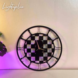 May include: Black and white chess-themed wall clock. The clock features a checkered center and a circular frame with chess piece designs. The clock hands are gold. The text "LightupLux" is in the upper left corner.
