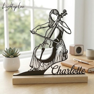 May include: A black metal cello player silhouette on a wooden base. The silhouette features a woman playing a cello, with the name "Charlotte" in cursive script. The top left corner has the text "LightupLux".