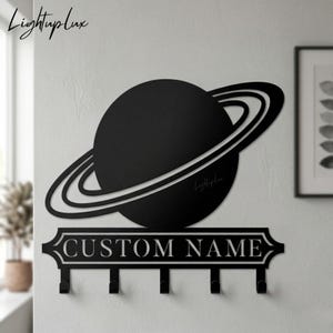 May include: Black metal wall-mounted key holder featuring a planet design with rings. The key holder has a rectangular section with the text "CUSTOM NAME" and four hooks below. The brand name "LightupLux" is also visible.
