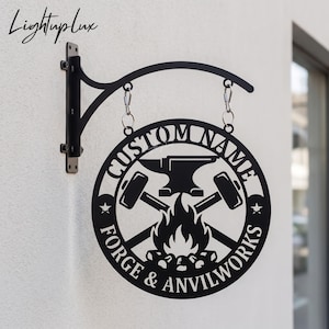May include: A black metal circular hanging sign featuring a cutout design of an anvil, crossed hammers, and a fire. The text reads CUSTOM NAME and FORGE & ANVILWORKS. The sign is suspended from a black metal wall-mounted bracket with two hooks.
