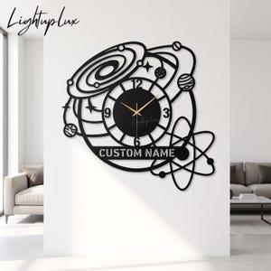 Könnte beinhalten: Eine schwarze Wanduhr mit einem Weltraum-Design, das Planeten, Sterne und eine Galaxie zeigt. Das runde Zifferblatt hat weiße Zahlen und goldene Zeiger. Ein horizontaler Balken am unteren Rand enthält den Text CUSTOM NAME in weißer Schrift.