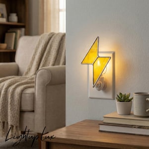 Può includere: Una luce notturna in vetro colorato giallo a forma di fulmine, inserita in una presa a muro. La luce emette un bagliore caldo, con una cornice metallica attorno ai segmenti di vetro. L'apparecchio è montato su una placca bianca.