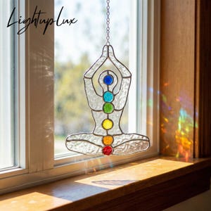 May include: A stained glass suncatcher shaped like a person in a seated yoga pose with hands raised. The figure is made of clear textured glass with six small circular glass accents in blue, turquoise, green, yellow, orange, and red.