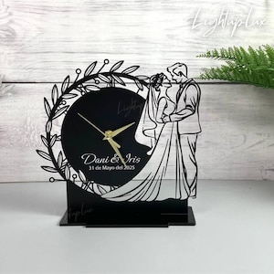 May include: Black acrylic wedding clock with a silhouette of a couple embracing, a clock face, and gold hands. The clock is decorated with a floral wreath and the names "Dani & Iris" and the date "31 de Mayo del 2025".