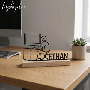 以下が含まれることがあります： 黒い金属製のデスクオーナメント。パソコンで作業する人を描いており、木製の台座に「ETHAN」という名前が黒い文字で書かれています。オーナメントは木製の机の上にあり、右側には鉢植えの多肉植物があります。「Lightuplux」というブランド名が左上にあります。