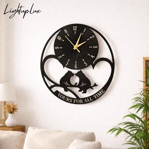 May include: A circular black wall clock featuring a central dial with gold hands and numerals. Below the dial, a cutout design depicts two bears sitting on a branch within a heart-shaped frame. The text YOURS FOR ALL TIME is inscribed at the bottom.