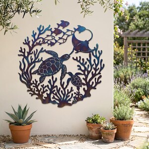 May include: A circular metal wall art piece featuring a sea turtle, a smaller turtle, a manta ray, and several fish swimming amidst coral reef structures. The metal has a gradient finish with blue, purple, and dark tones throughout the design.