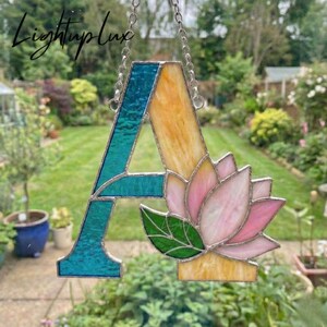 May include: A stained glass letter A suncatcher featuring a textured teal left side and a smooth yellow right side. A pink lotus flower with a green leaf is attached to the lower right section of the letter, suspended by a silver metal chain.