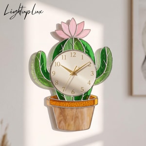 May include: A wall clock shaped like a green cactus in a tan pot with a pink flower on top. The clock face is circular with gold numbers and hands. The design features stained glass style panels with silver metal outlines and the text Lightuplux.