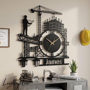 May include: A black metal wall clock featuring a construction theme with a crane, a building frame, and a silhouette of a person holding blueprints. The clock face is circular with gold hands and the name James is cut out at the bottom of the design.