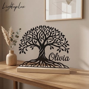 May include: Black metal tree of life sculpture with the name "Olivia" on a light wood base. The tree has detailed branches and roots. The text "Lightuplux" is in the upper left corner. The sculpture is on a wooden table.
