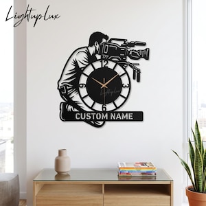 Op de afbeelding: Een zwarte wandklok met het silhouet van een persoon die een professionele videocamera bedient. De wijzerplaat heeft witte cijfers en dunne gouden wijzers. Onder de klok staat een balk met de tekst CUSTOM NAME.