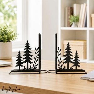 May include: A pair of black metal bookends featuring a silhouette design of evergreen pine trees and leafy branches. Each bookend has a flat base and a vertical panel with a cut-out forest motif, providing a stable support for books.