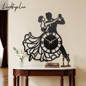 Puede incluir: Un reloj de pared negro con la silueta de una pareja bailando. La esfera del reloj está integrada en el vestido fluido de la mujer, mostrando manecillas y números dorados en las posiciones 3, 6, 9 y 12. El texto Lightuplux es visible arriba a la izquierda.