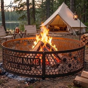 May include: A circular metal fire pit ring featuring a laser-cut honeycomb pattern, sunflower motifs, and bees. The metal panels display the text CUSTOM NAME and EST. 2026. A bright fire burns inside the ring, which is set in a wooded campsite.