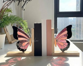 May include: A pair of wooden bookends featuring stained glass butterfly wings in shades of pink and black. The wings are attached to light-colored wooden bases, supporting a black book and a white book between them on a flat surface.
