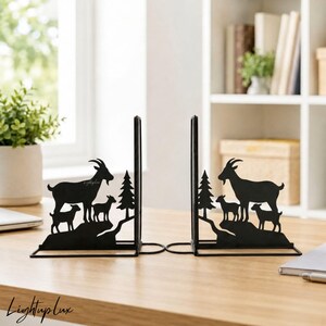 May include: A pair of black metal bookends featuring a silhouette design of a mountain goat with two kids standing on a rocky ledge next to a pine tree. The metal cutouts are flat and display a detailed outdoor scene with animals in nature.