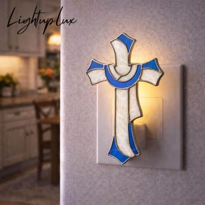 May include: A plug-in night light shaped like a cross, featuring white and blue stained glass panels with a metallic frame. The light emits a warm glow from behind the cross, which is mounted on a standard white wall outlet cover.