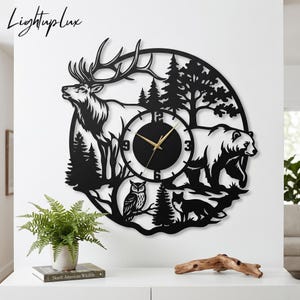 Könnte beinhalten: Eine kreisförmige schwarze Wanduhr mit einer lasergeschnittenen Silhouette einer Waldszene mit einem Elch, einem Bären, einer Eule und einem Fuchs. Das Zifferblatt ist mittig im Design platziert und zeigt schwarze Ziffern bei 3, 6, 9 und 12 sowie goldene Zeiger.