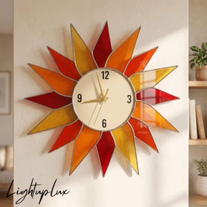 May include: A sunburst wall clock featuring a circular cream-colored face with black numerals at 3, 6, 9, and 12 o'clock. The clock is surrounded by pointed, petal-like rays in alternating shades of translucent red, orange, and yellow glass.