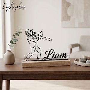 Op de afbeelding: Een zwart metalen silhouet van een persoon die trombone speelt, gemonteerd op een rechthoekige houten voet. Het ontwerp bevat de naam Liam in een cursief lettertype. Het object staat op een houten oppervlak naast een vaas.