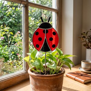 Pode incluir: Um ornamento de vitral em forma de joaninha, com asas vermelho-brilhantes com pintas pretas e cabeça preta, montado numa haste metálica fina. A joaninha está posicionada num vaso de terracota com uma planta verde junto a uma janela.