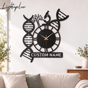 Puede incluir: Un reloj de pared negro con esfera circular, números romanos y manecillas doradas. El diseño incluye siluetas recortadas de dos cadenas de ADN, un árbol y un microscopio. Debajo de la esfera hay una placa rectangular con el texto CUSTOM NAME.