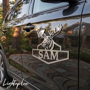 May include: A metallic silver car decal featuring a stylised moose head silhouette positioned above a geometric frame containing the name SAM in bold capital letters. The emblem is adhered to the side door of a dark grey vehicle.