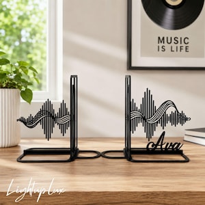 May include: A pair of black metal bookends featuring a stylized sound wave design. The right bookend includes the name Ava in a cursive script integrated into the sound wave pattern. The bookends have a flat base and vertical support structure.