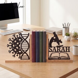 May include: A pair of wooden bookends featuring black metal cutouts. The left piece displays a virus shape and a DNA double helix. The right piece features a microscope, an open book, and the name SARAH in bold, capitalized letters.