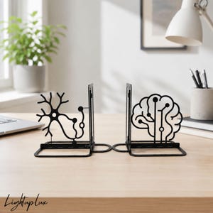 Op de afbeelding: Een paar boekensteunen van zwart metaal met wetenschappelijke silhouetten. De ene boekensteun toont een gedetailleerde neuronstructuur met vertakte dendrieten, de andere een menselijk brein geïntegreerd met printplaatlijnen.