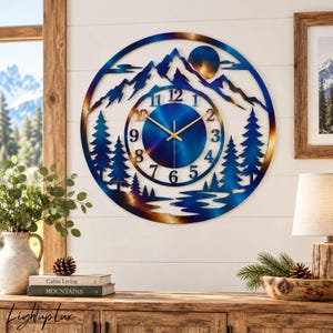 May include: A circular metal wall clock featuring a mountain landscape with pine trees and a river. The design has a blue and bronze gradient finish. The clock face has gold-colored hands and black numerals from 1 to 12 arranged in a circle.