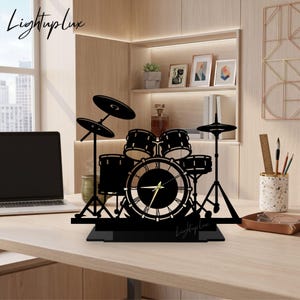Puede incluir: Un reloj de escritorio negro cortado con láser en forma de batería, con una esfera circular central y manecillas doradas. El diseño incluye siluetas de varios tambores y platillos sobre soportes, con la marca Lightuplux visible.