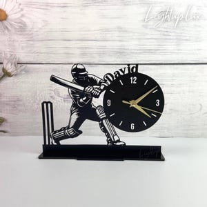 May include: A black desk clock featuring a silhouette of a cricket batter in a batting stance next to a set of wickets. The clock face is circular with gold hands and the name David printed in a script font above the numbers 12, 3, 6, and 9.