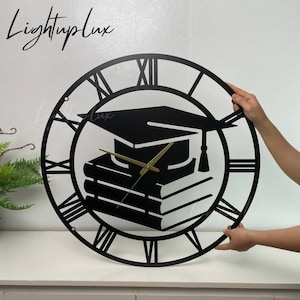 May include: Black metal wall clock with Roman numeral markings. The clock face features a graduation cap and stack of books.