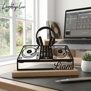 Op de afbeelding: Zwart metalen silhouet van een DJ-controller en koptelefoon op een houten voet, met de naam "Liam" gegraveerd. Het ontwerp staat op een bureau met een laptop en een kleine plant in een pot. Het logo "LightupLux" staat in de linkerbovenhoek.
