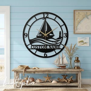May include: A circular black metal wall clock featuring a cutout design of a sailboat on waves and a lighthouse. The clock face includes the text CUSTOM NAME, large numerals at the 3, 6, 9, and 12 positions, and thin gold-colored clock hands.