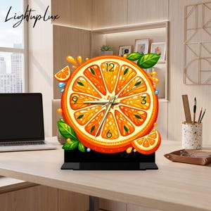 Könnte beinhalten: Eine runde Tischuhr in Form einer Orangenscheibe mit leuchtend orangen Segmenten, grünen Blättern und Wassertropfen. Das Zifferblatt zeigt schwarze Zahlen an den Positionen 12, 3, 6 und 9 mit goldfarbenen Zeigern auf einem schwarzen Sockel.