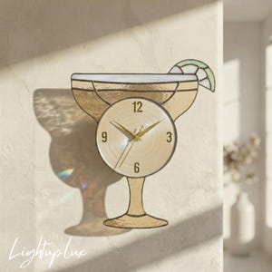 Puede incluir: Un reloj de pared de vitral con forma de copa de margarita en tono beige claro. La esfera circular presenta números dorados 3, 6, 9 y 12 con manecillas doradas a juego. Un pequeño gajo de lima de vitral verde se encuentra en el borde.
