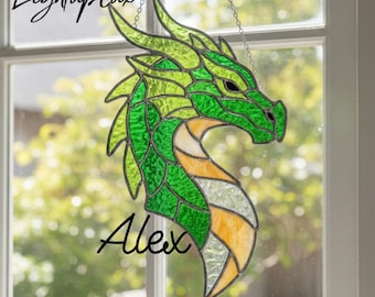 May include: A stained glass dragon suncatcher featuring shades of green, yellow, and clear textured glass with dark metal soldering. The piece includes the text LightupLux at the top and the name Alex in black script at the bottom.