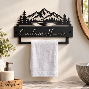 May include: A black metal wall-mounted towel rack featuring a laser-cut silhouette of a mountain range and pine trees above a rectangular nameplate with the text Custom Name, holding a folded white towel on the lower horizontal bar.