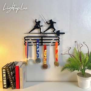 May include: Black metal fencing medal holder with silhouettes of fencers and the name "JAMES". The holder displays several medals with colorful ribbons. The logo "LightupLux" is in the upper left corner.