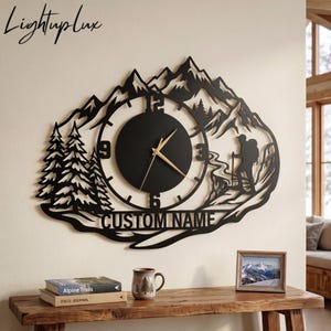 May include: A black metal wall clock featuring a mountain range silhouette, pine trees, and a hiker with a walking stick. The circular clock face has gold hands and numbers 3, 6, 9, and 12. The text CUSTOM NAME is cut out at the bottom of the design.