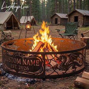 May include: A circular metal fire pit ring featuring laser-cut dragon silhouettes and the text CUSTOM NAME EST. 2026. The dark brown metal structure surrounds a bright, active campfire with burning logs, set in a wooded outdoor campsite.