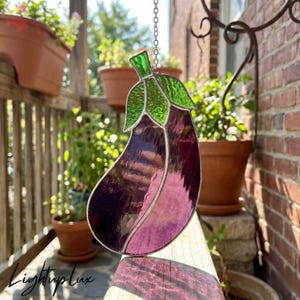 May include: Stained glass eggplant suncatcher with a chain for hanging. The eggplant is a mix of purple and pink glass, with green leaves. The suncatcher is hanging on a porch with plants in the background.