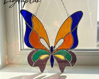 May include: A stained glass butterfly suncatcher featuring vibrant segments of blue, orange, yellow, green, and purple glass held together by dark metal solder lines. The butterfly is suspended by a thin silver-colored metal chain for hanging.